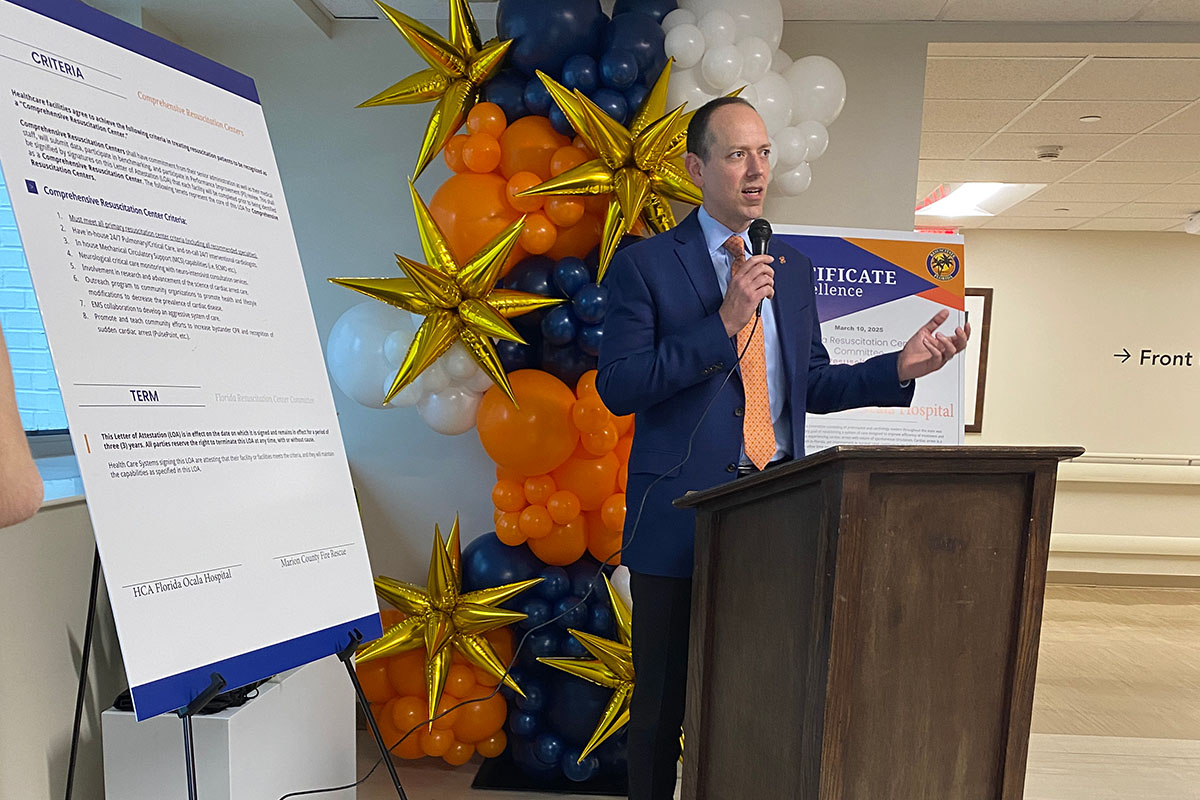 A man in a blue suit and orange tie giving a speech while holding a microphone with an informational board behind him