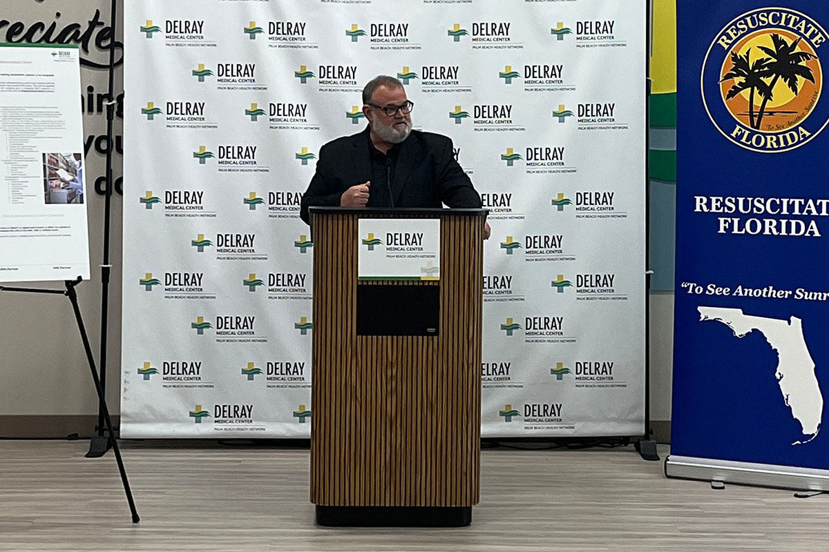 A man in a black suit at a podium giving a speech with a Resuscitate Florida banner beside him