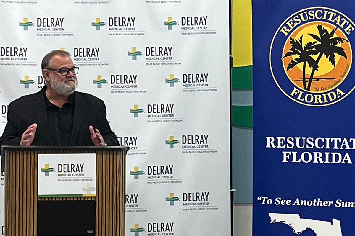 A man in a black suit at a podium with a Florida Resuscitate banner beside him